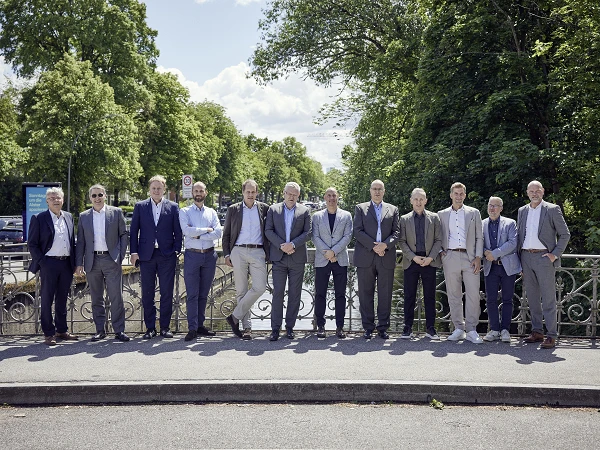 Eine Gruppe von zwölf Männern in Anzügen steht auf einer Brücke, umgeben von Bäumen und einem klaren Himmel im Hintergrund. Sie sind in einem entspannten, freundlichen Arrangement aufgestellt.