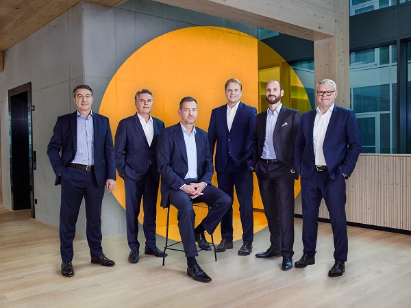Sechs Männer in Anzügen stehen und sitzen in einem modernen Büroumfeld vor einer großen, orangefarbenen Wandinstallation. Sie posieren in einer Reihe, während sie in die Kamera schauen.