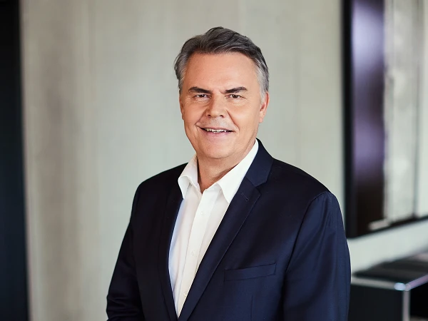 A man with gray hair is smiling, dressed in a dark blazer and white shirt, standing in a modern interior space.