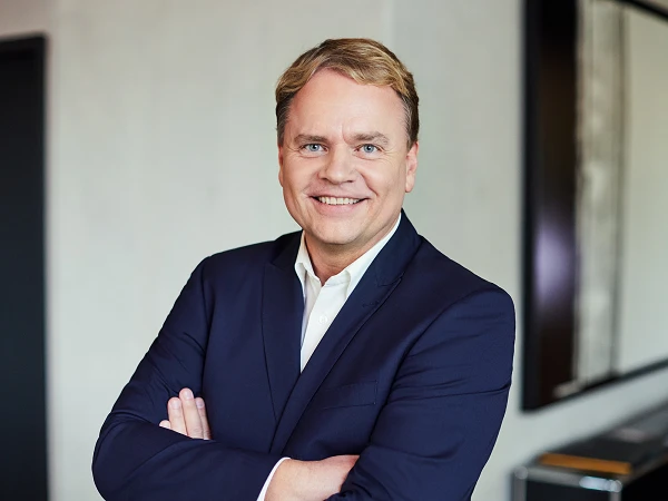 Ein gut gekleideter Mann mit blonden, kurzen Haaren lächelt freundlich in die Kamera, während er mit verschränkten Armen vor einem hellen, modernen Hintergrund steht.
