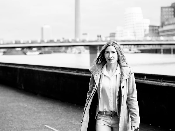 Frau im Mantel geht lächelnd am Rhein entlang, moderne Architektur im Hintergrund
