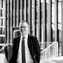 Mann mit Brille im Anzug vor Bürogebäude, freundlich blickend