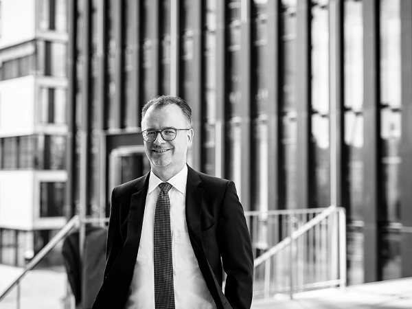 Mann mit Brille im Anzug vor Bürogebäude, freundlich blickend