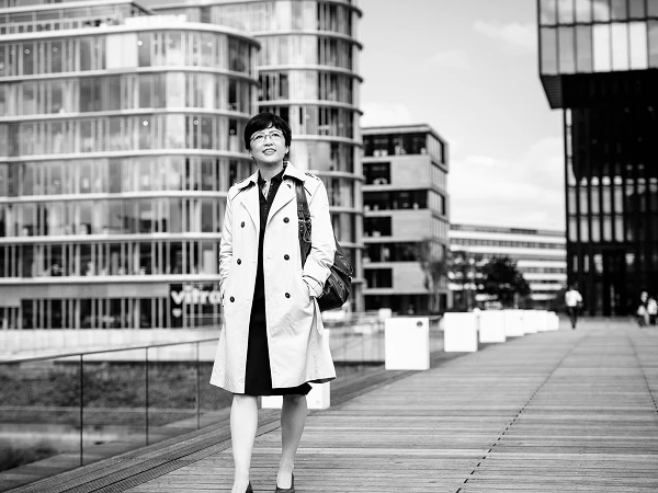 Frau im Trenchcoat auf Brücke vor moderner Architektur, freundlich blickend