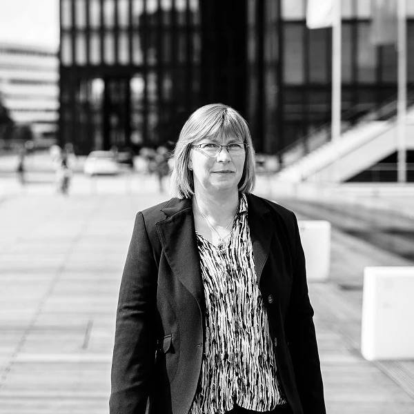 Frau mit Brille im Blazer vor urbanem Hintergrund, leicht lächelnd