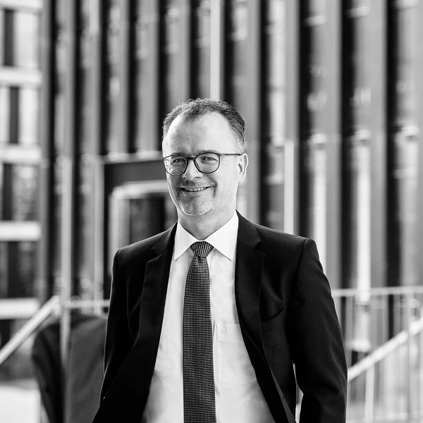 Man in suit with glasses, smiling warmly in front of modern architecture
